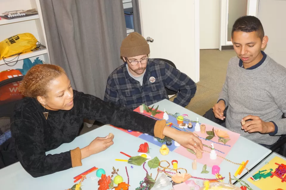 Three people gather around a table to discuss maps made of toys and found objects.